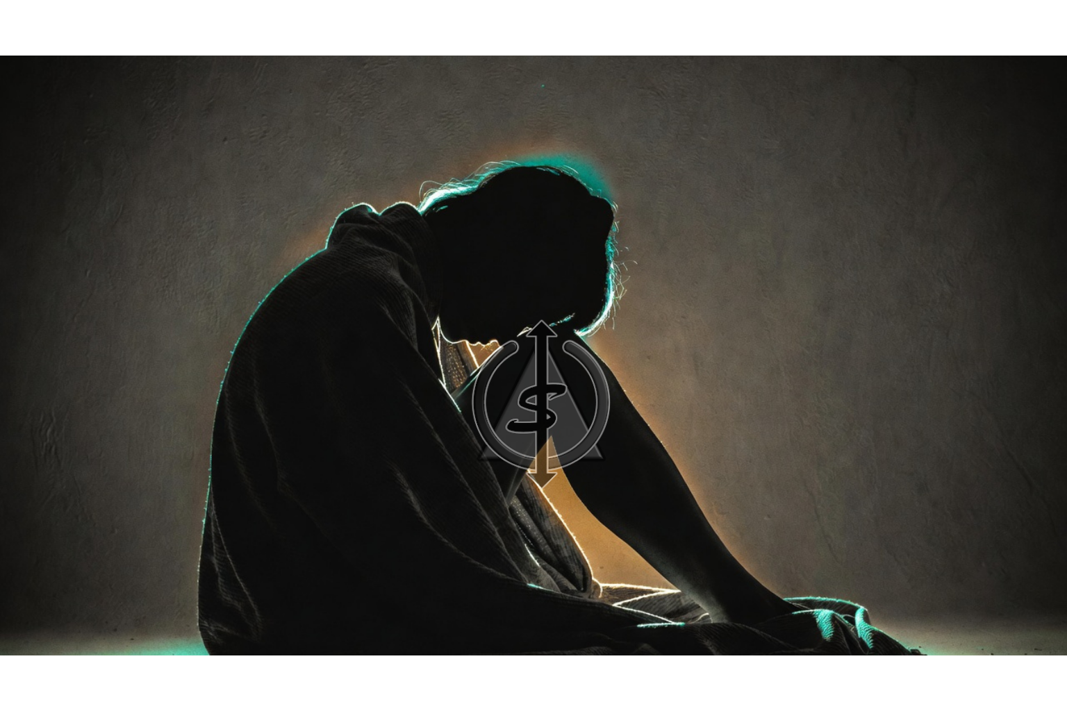 Black and white image of a person sitting wrapped in a soft blanket, resting against a wall with subtle teal and gold light outlining their silhouette.