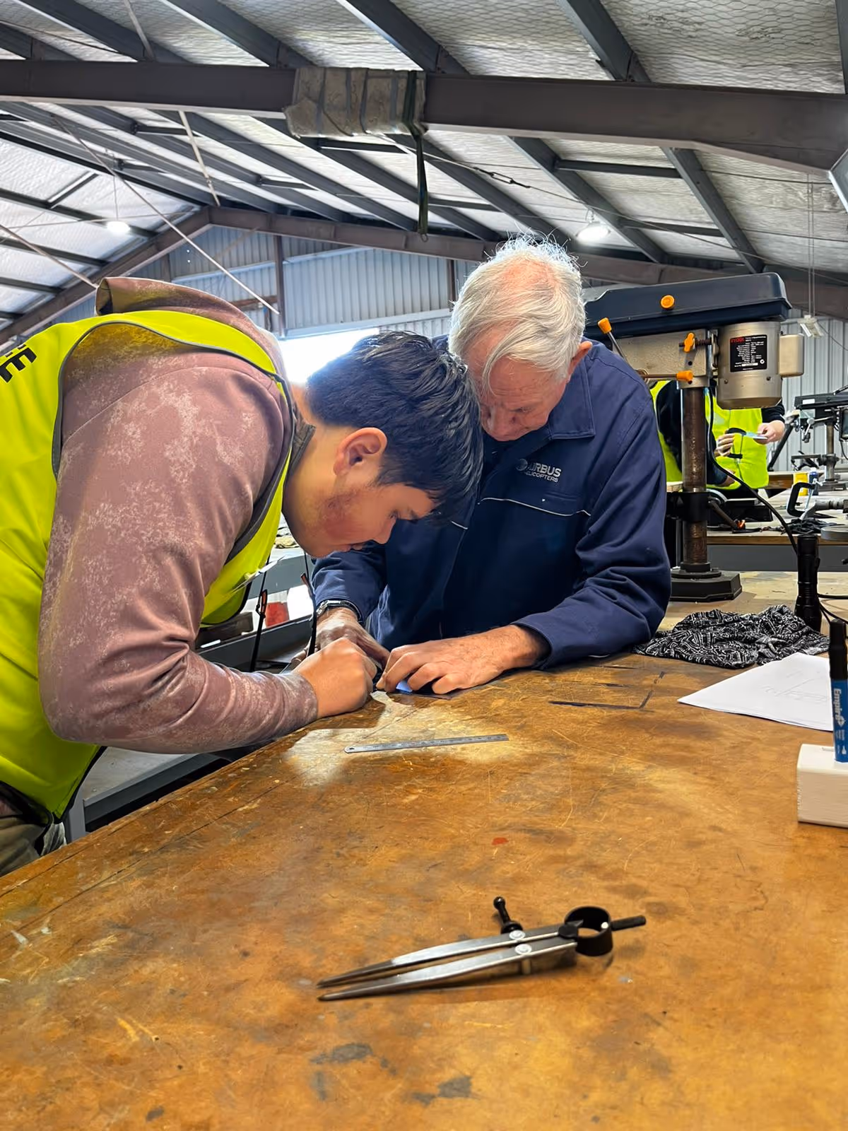 Two men closely working together at a wooden workbench inside a workshop, focusing on a small project with tools and papers around.