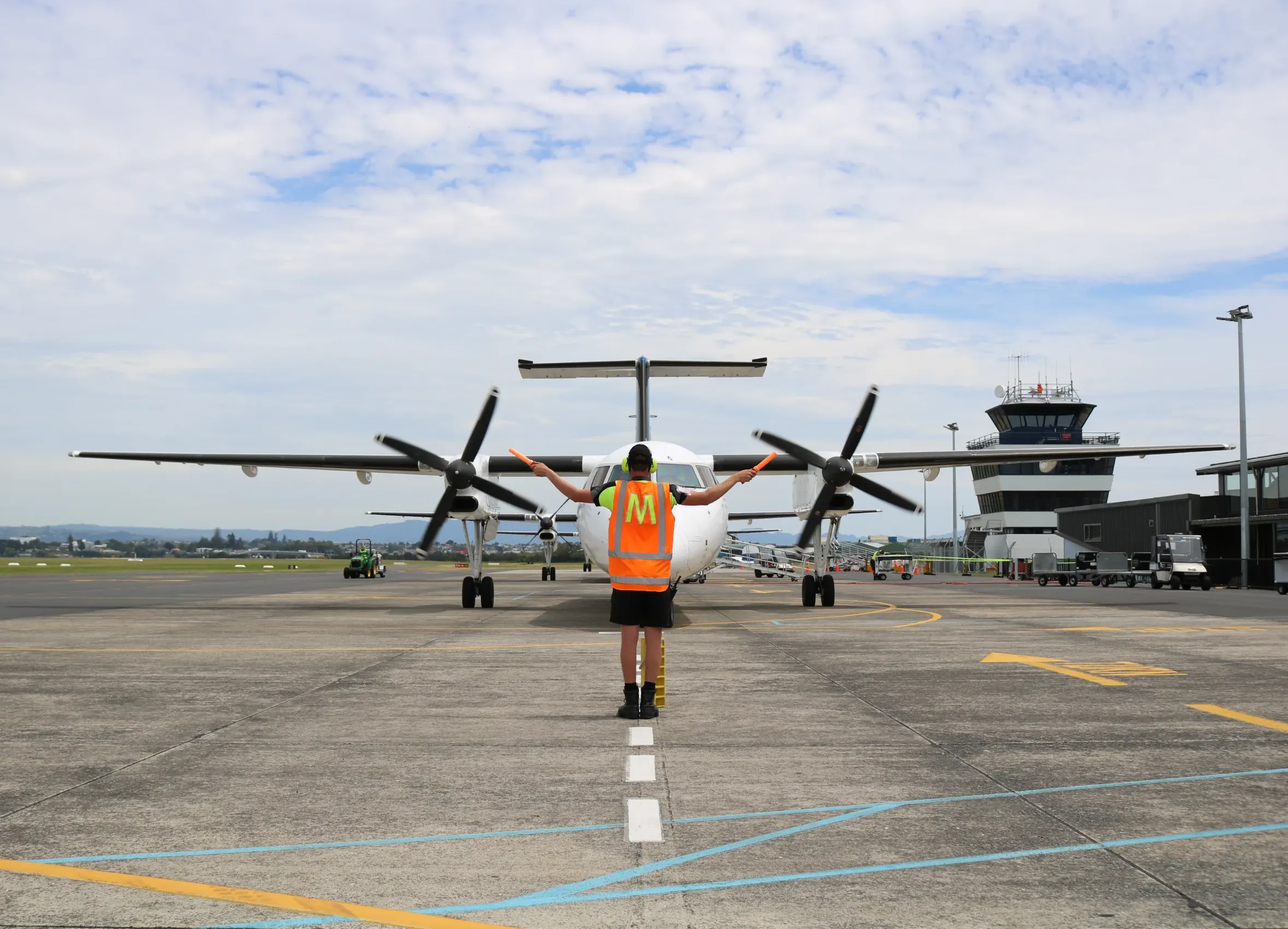 Airport ground staff in orange vest guiding a propeller airplane on the tarmac with control tower in the background.
