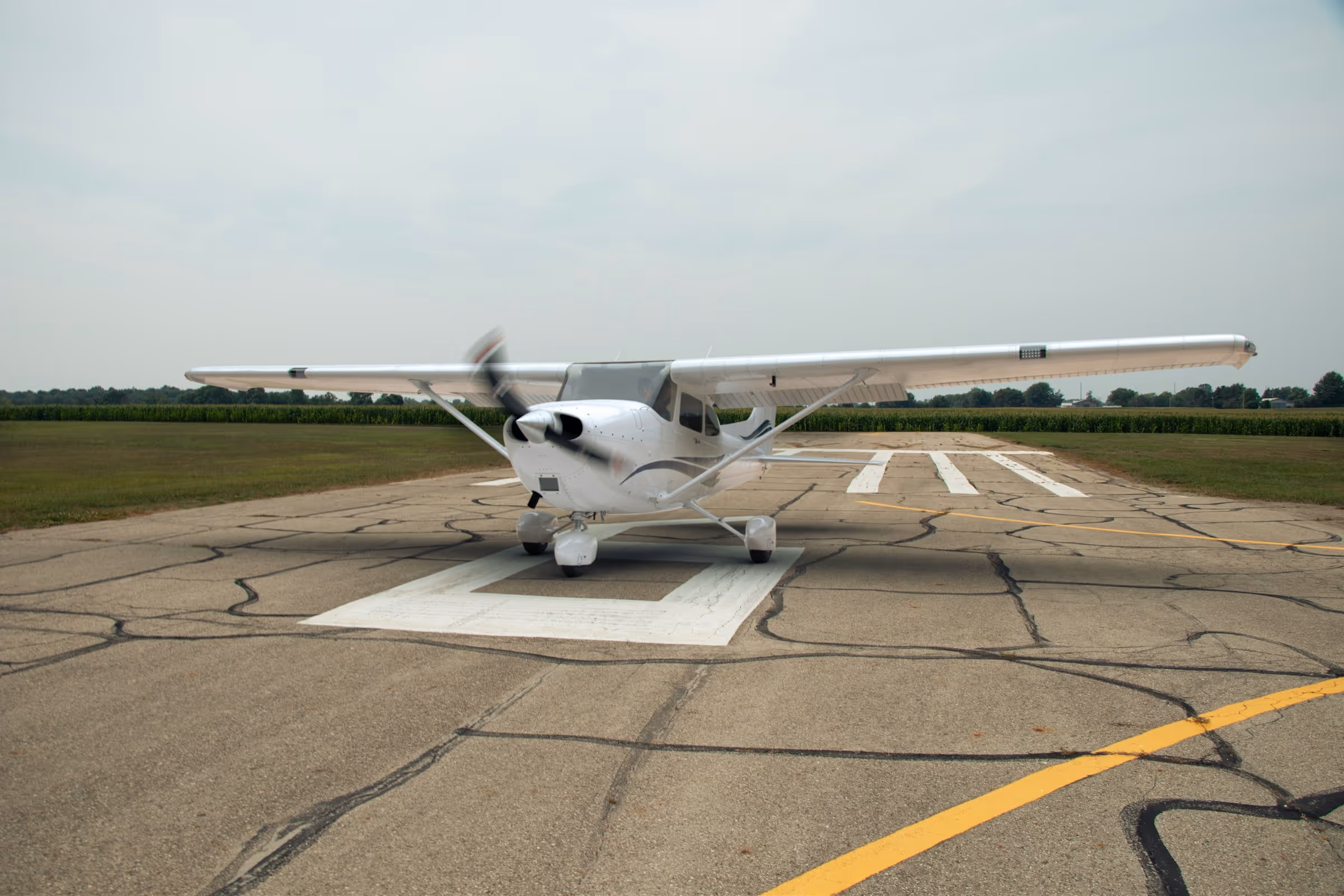 White small single-engine airplane on a cracked runway with grass and trees in the background.