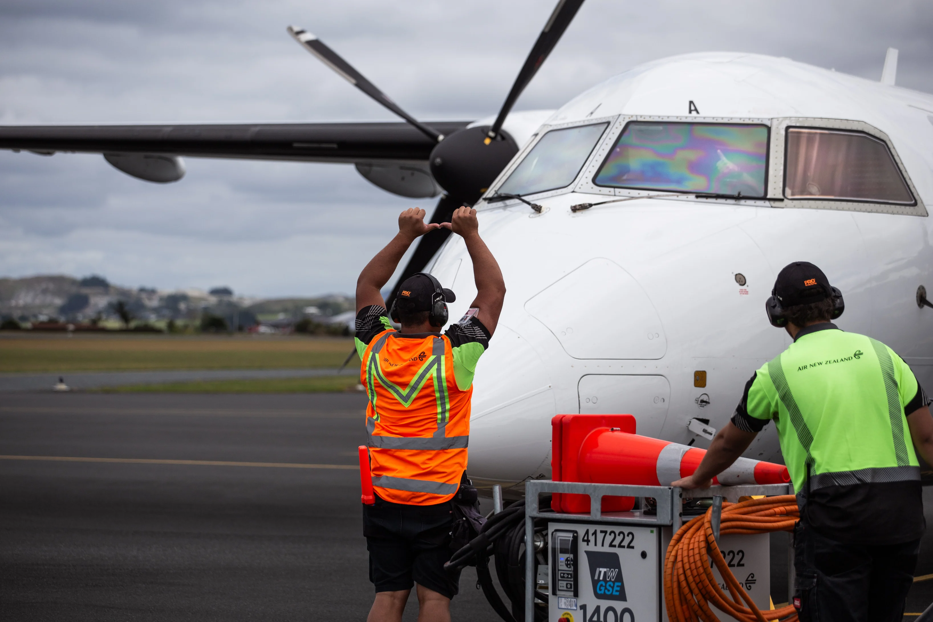 Two ground crew members in Air New Zealand uniforms preparing a propeller airplane on the runway with one directing the plane using hand signals.
