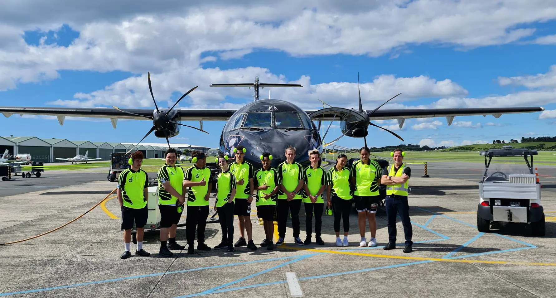 Group of people in bright green shirts standing in front of a black twin-propeller airplane on an airport tarmac under a blue sky with clouds.