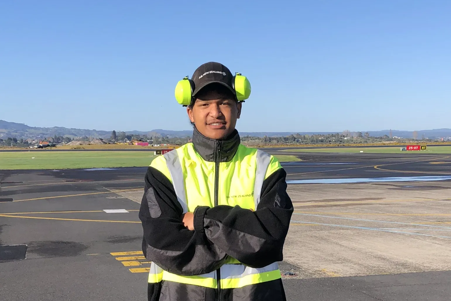 Man in high-visibility jacket and hearing protection standing on an airport runway with hills in the background.