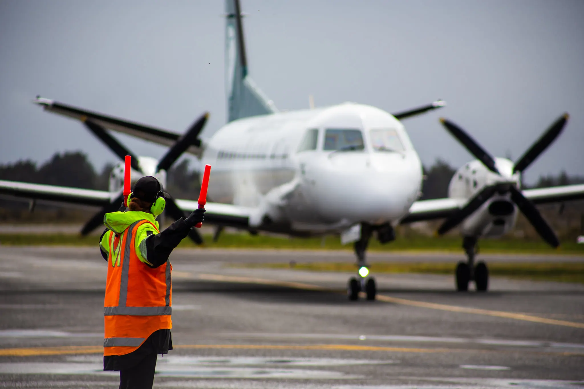 Airport ground crew member in orange safety vest signaling with orange wands to a white propeller airplane taxiing on runway.