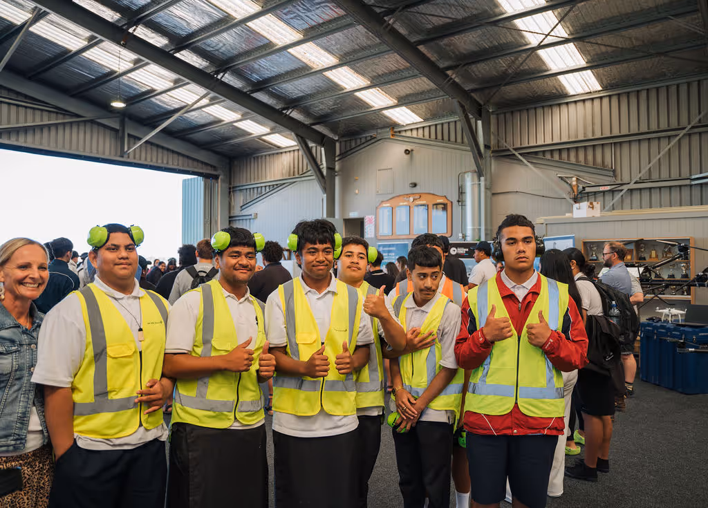 Group of young men wearing yellow safety vests and green ear protection posing with thumbs up inside a large industrial building.