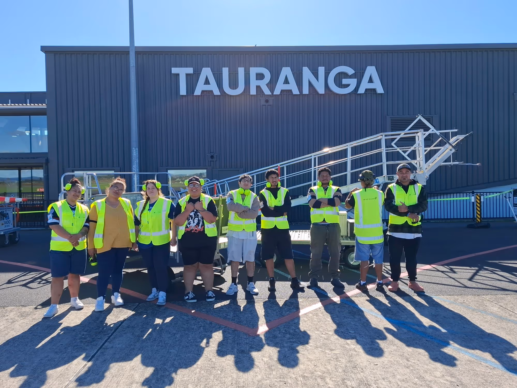 Group of people wearing yellow safety vests and ear protection standing in front of a building with the sign 'TAURANGA' on a sunny day.