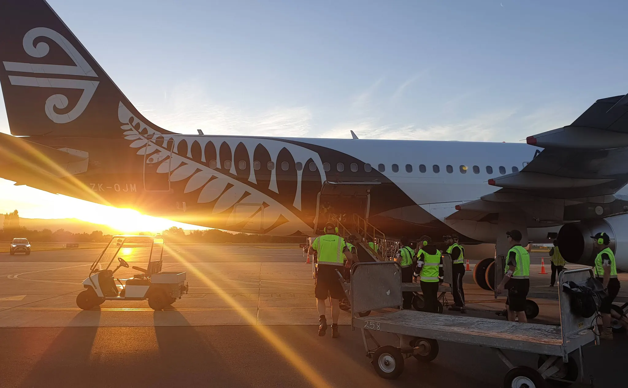 Ground crew wearing neon vests loading luggage into an Air New Zealand airplane at sunset.