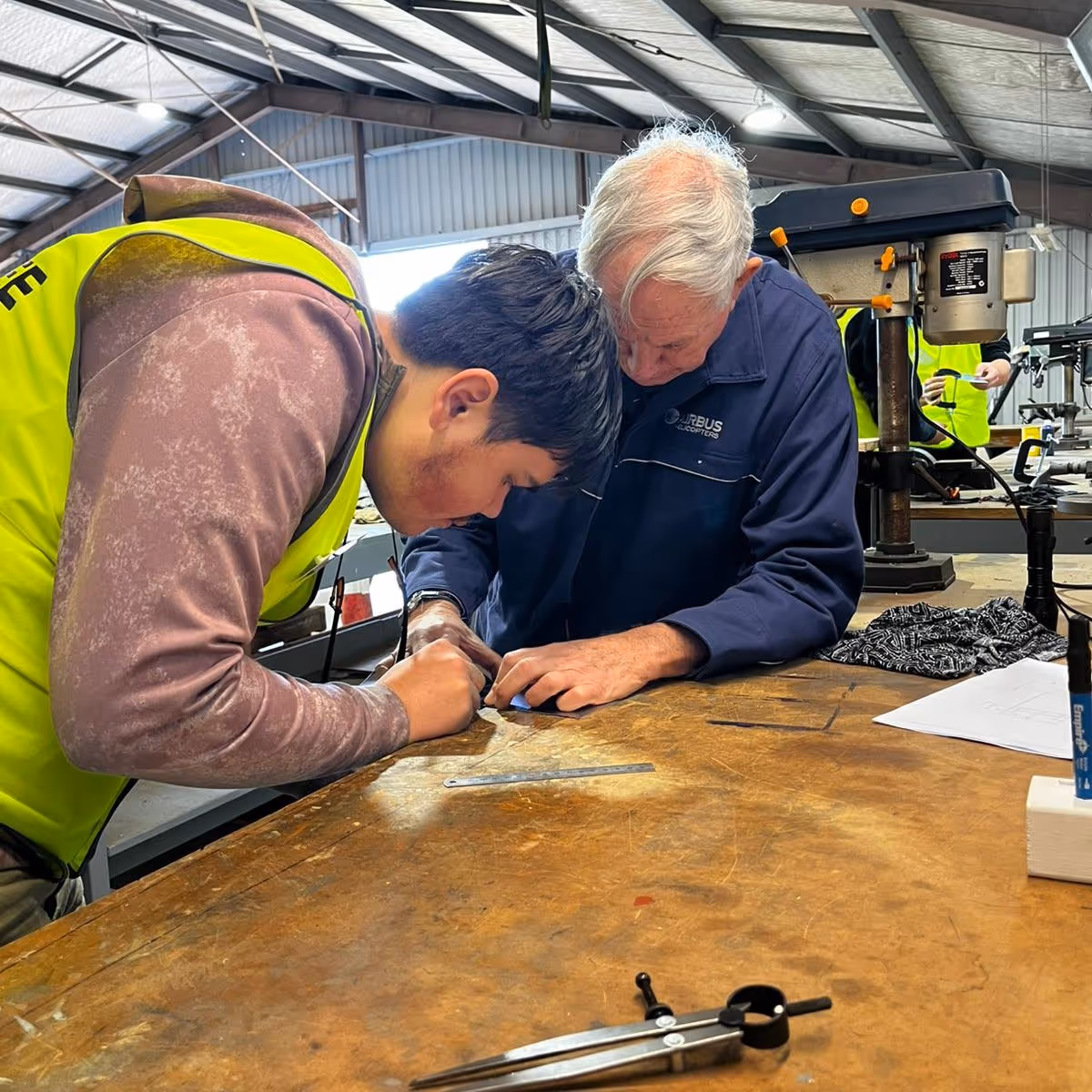Two men closely examining and working on a small object on a wooden workbench in a workshop.