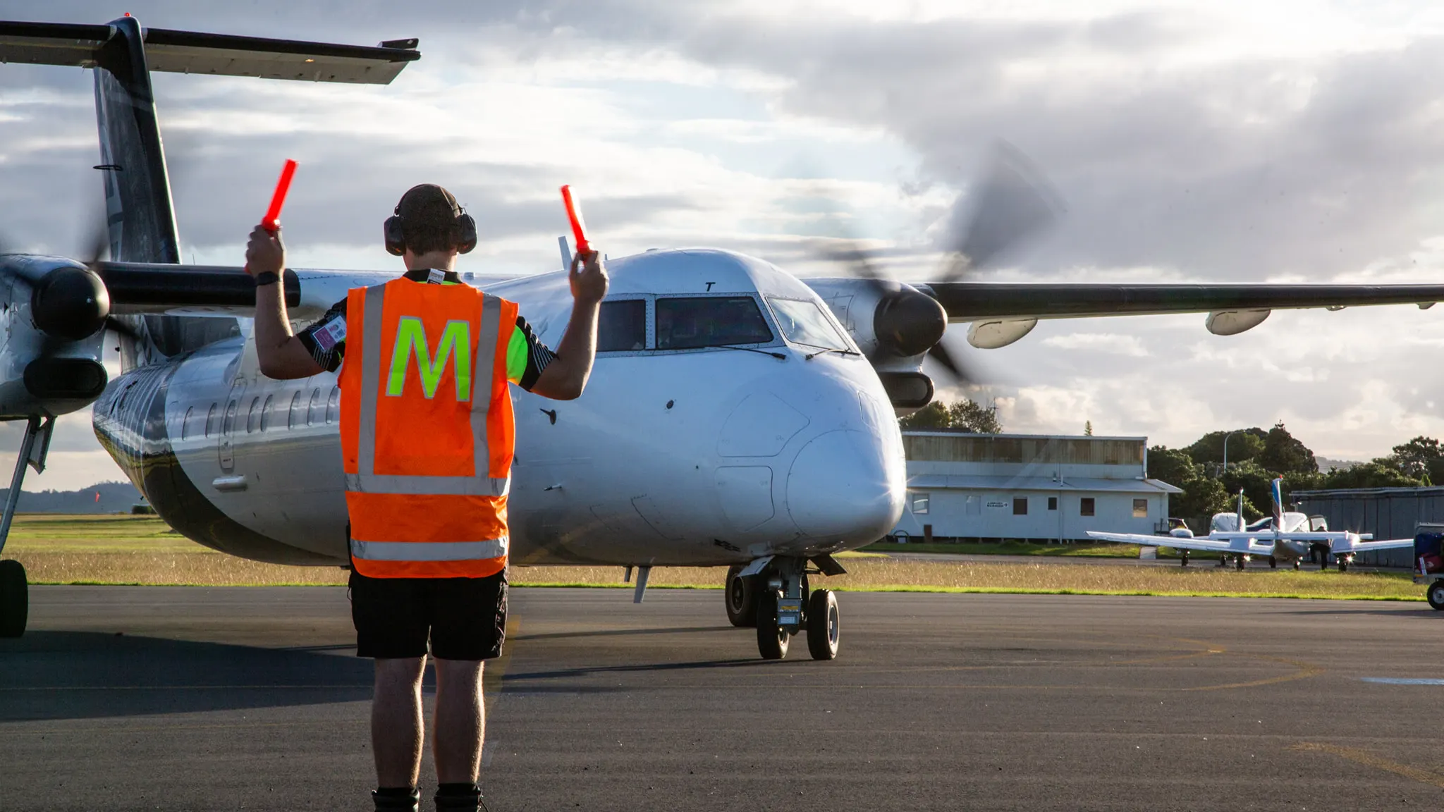 Airport ground crew member in orange safety vest directing a white twin-propeller aircraft on the tarmac.