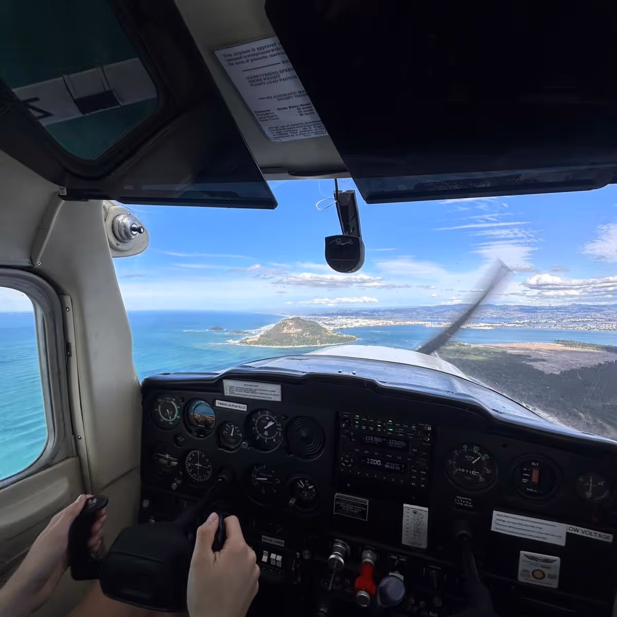 Pilot's hands on controls inside small airplane cockpit flying over coastline with ocean and hills.