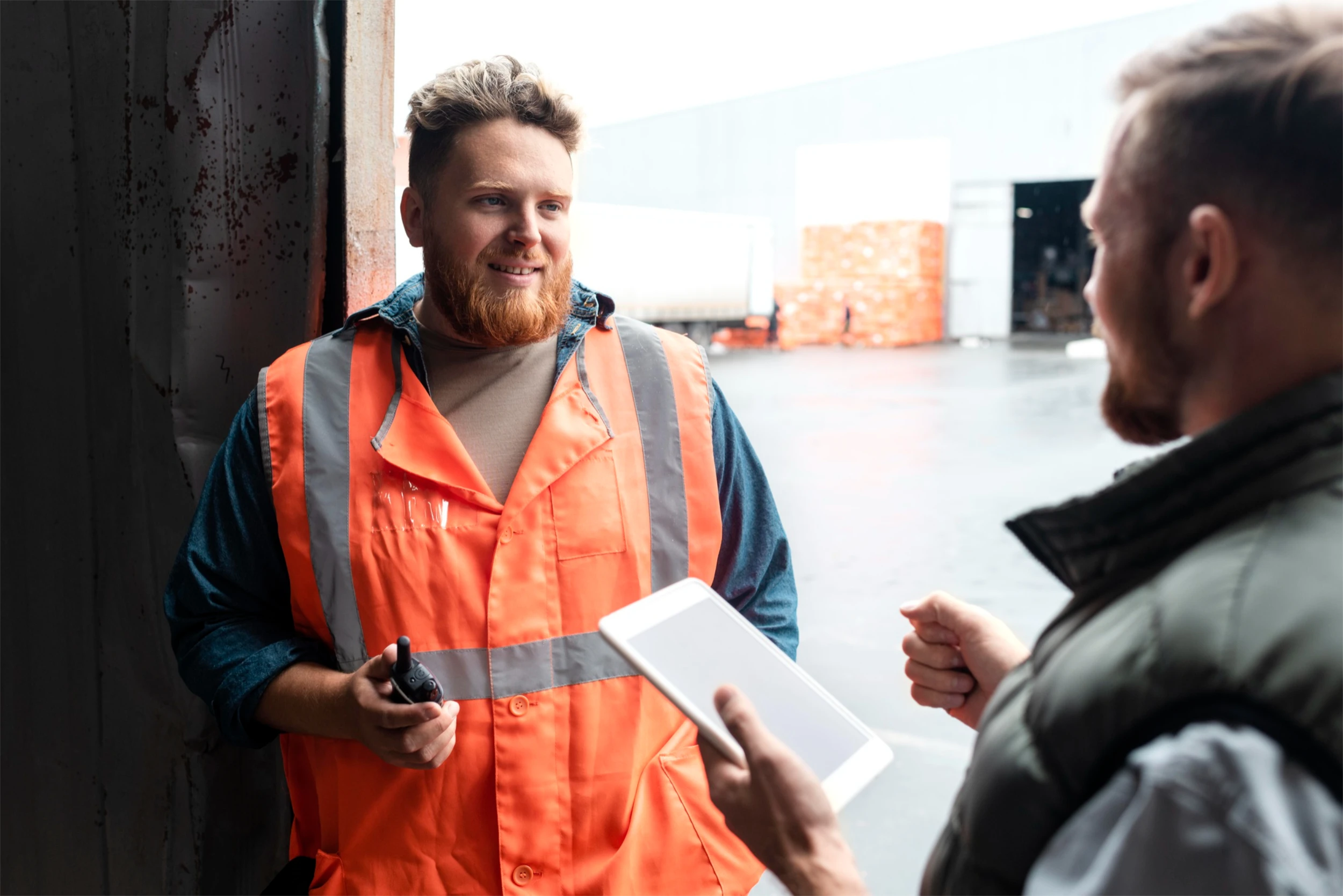 Two warehouse workers talking, one wearing an orange safety vest holding a walkie-talkie, the other holding a white tablet.