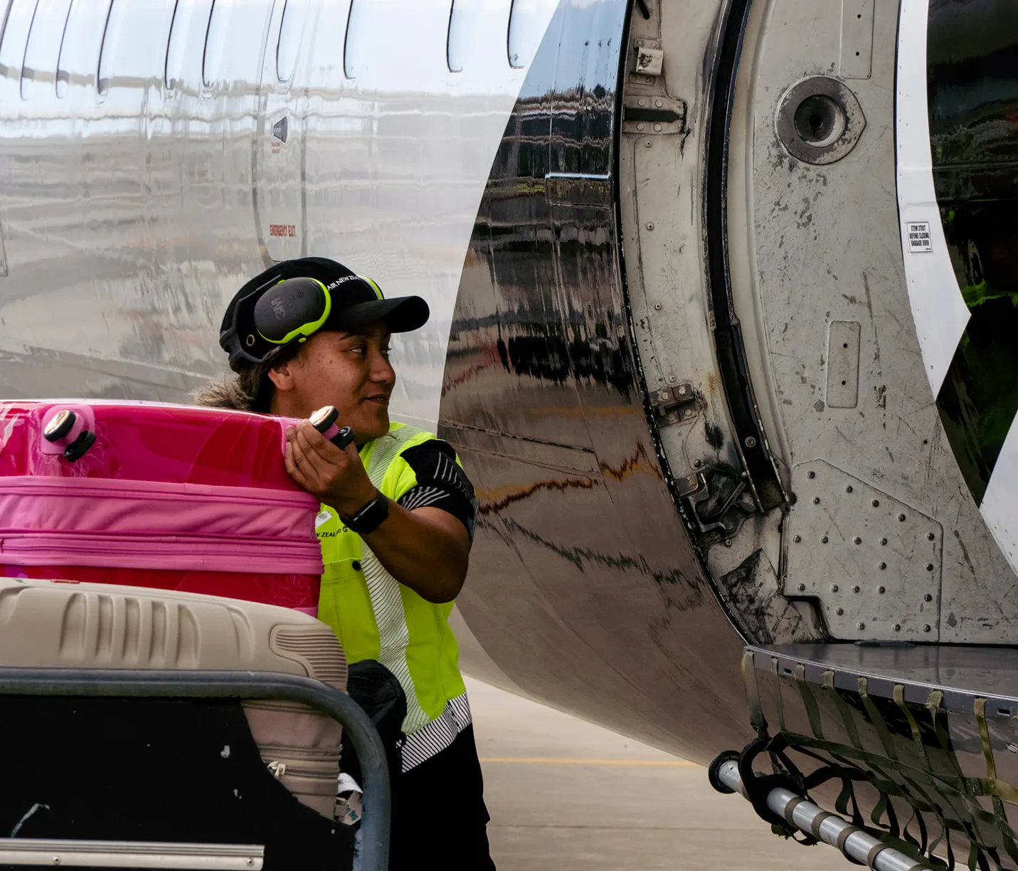 Airport worker in high-visibility vest loading pink and beige suitcases into an airplane cargo hold.