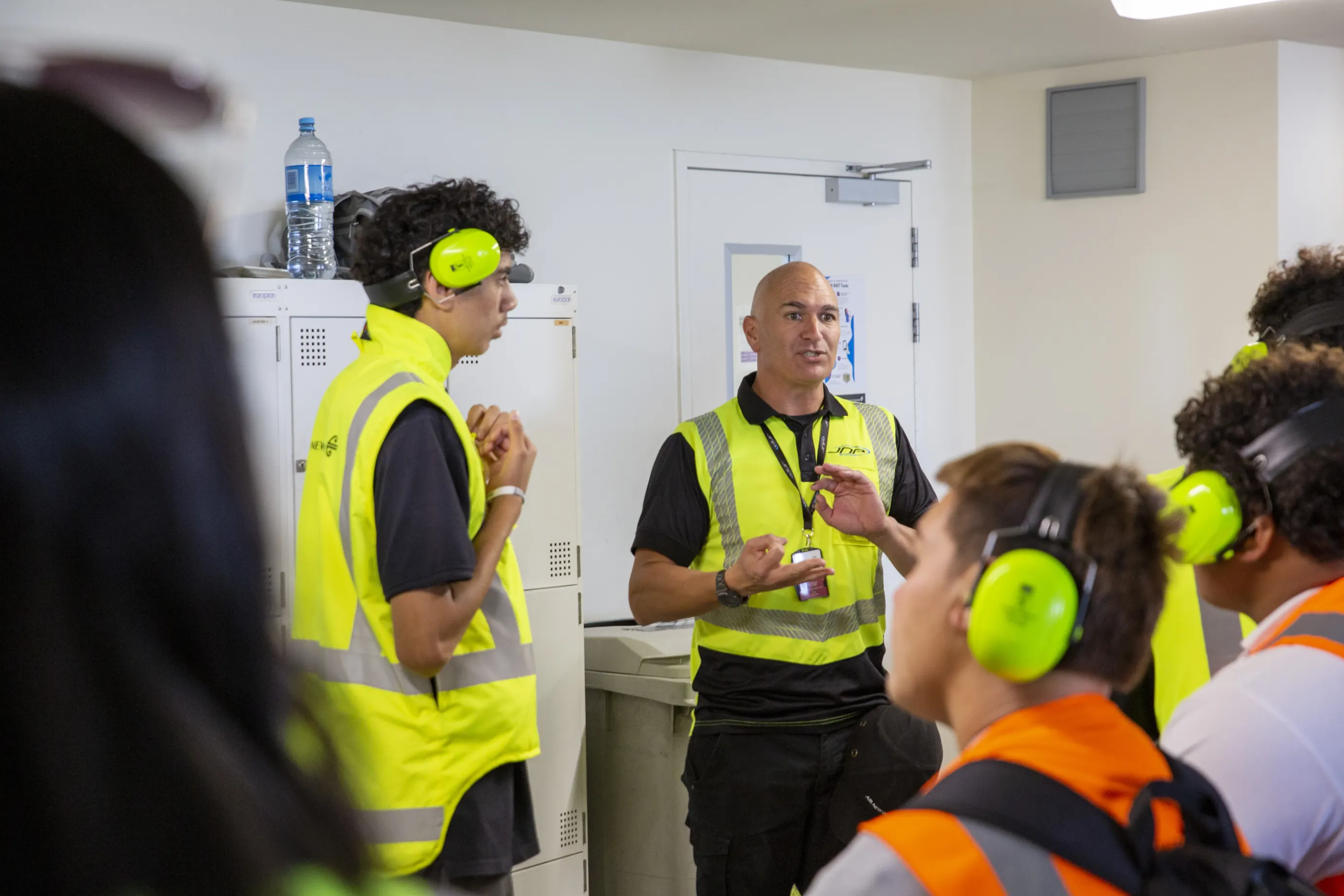 Safety briefing in progress with a man in a yellow reflective vest speaking to a group wearing safety headphones and vests.