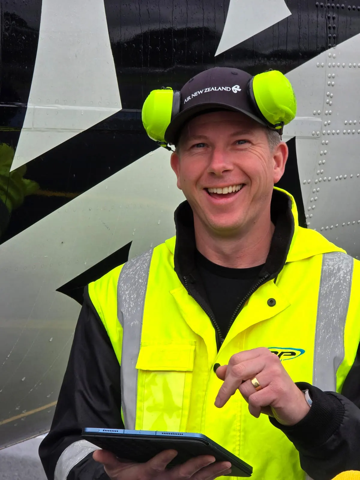 Smiling man in high-visibility jacket and Air New Zealand cap holding a tablet in front of an airplane.