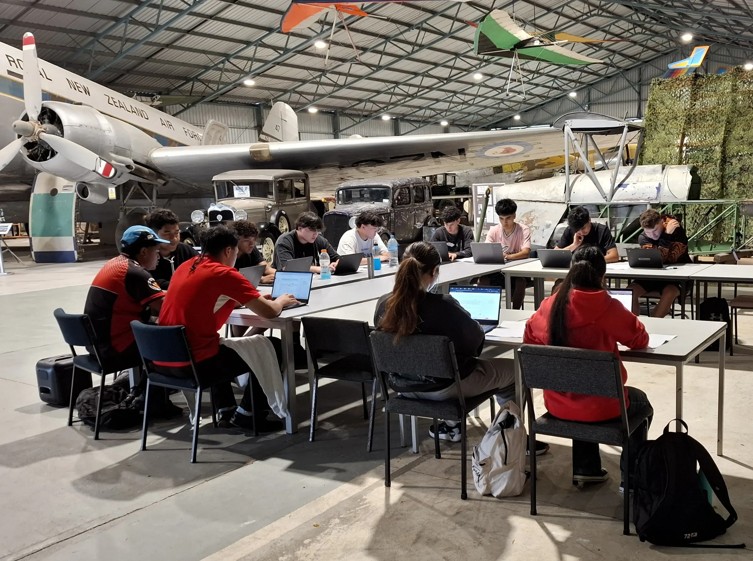 Group of young people sitting at tables with laptops inside an aircraft hangar, with vintage planes and cars in the background.