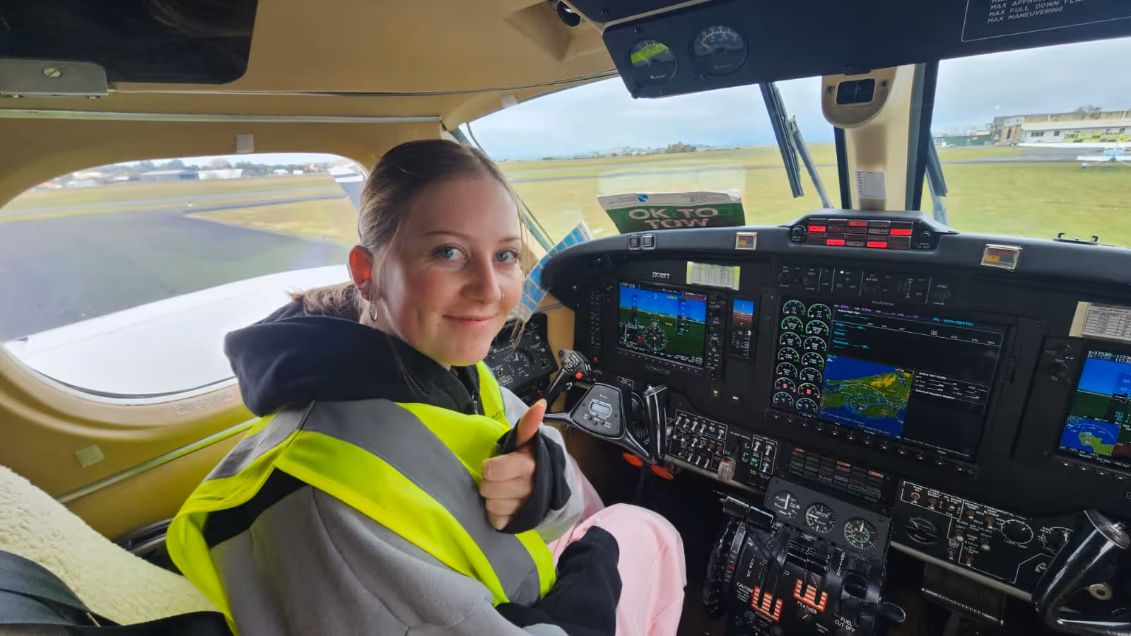 Woman wearing a high-visibility vest gives a thumbs up while sitting in the cockpit of an airplane.