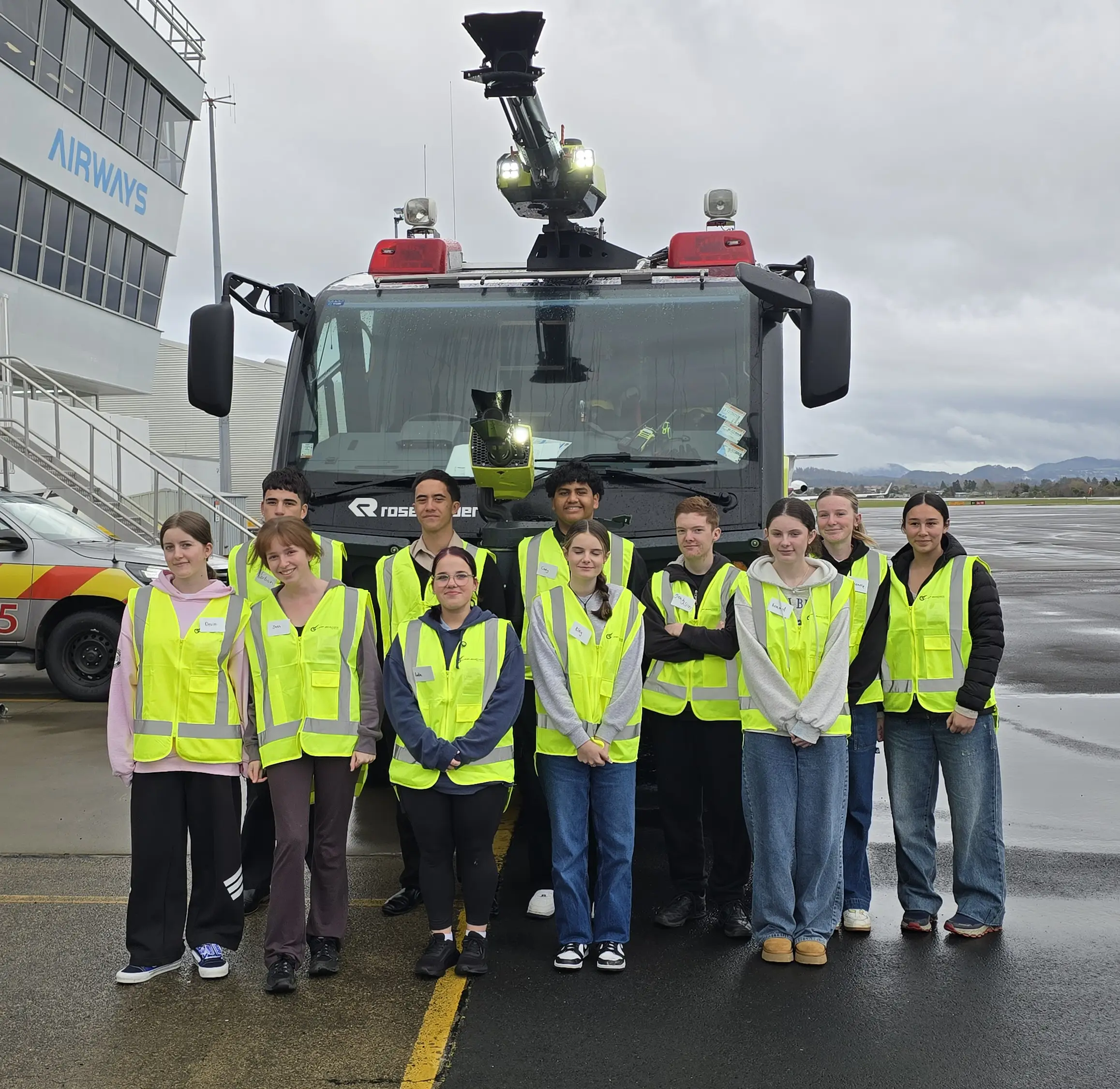 Group of young people wearing yellow safety vests standing in front of a fire truck at an airport under cloudy sky.
