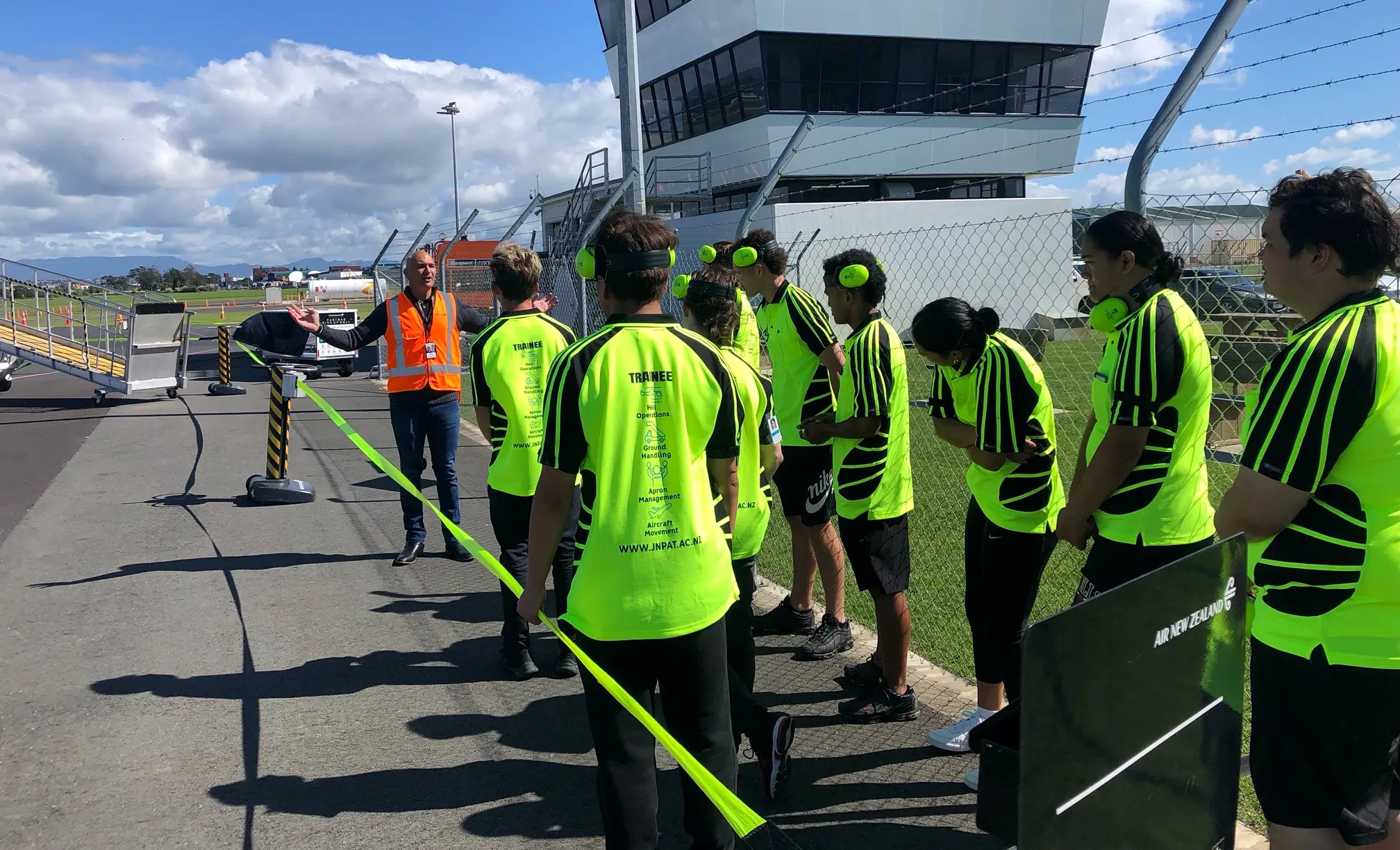 Instructor in orange safety vest explaining safety procedures to a group of trainees in neon yellow shirts with ear protection at an airport ramp.