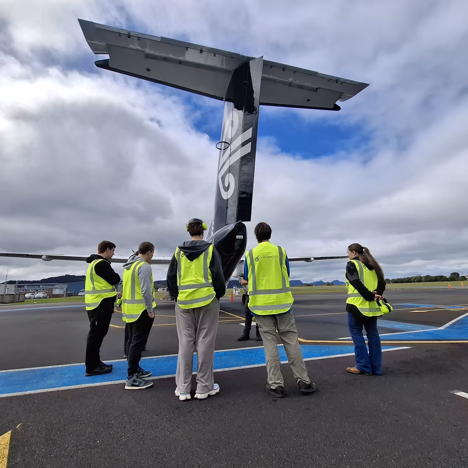 Five people wearing yellow safety vests standing on an airport tarmac near the tail of an Air New Zealand airplane.