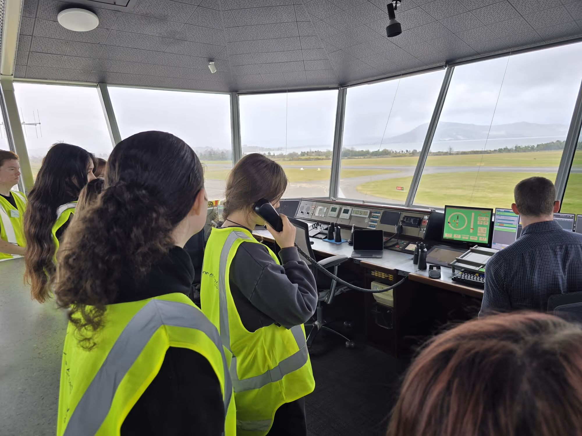 Group of people wearing yellow safety vests inside an air traffic control tower, one person speaking on a phone while others observe.