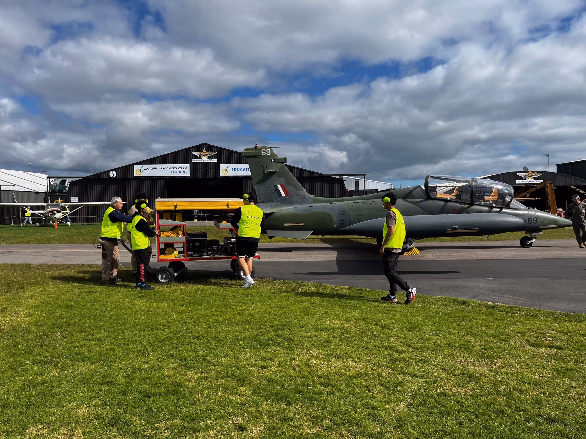 Five people wearing yellow safety vests and ear protection near a military training jet parked on an airfield with hangars in the background.