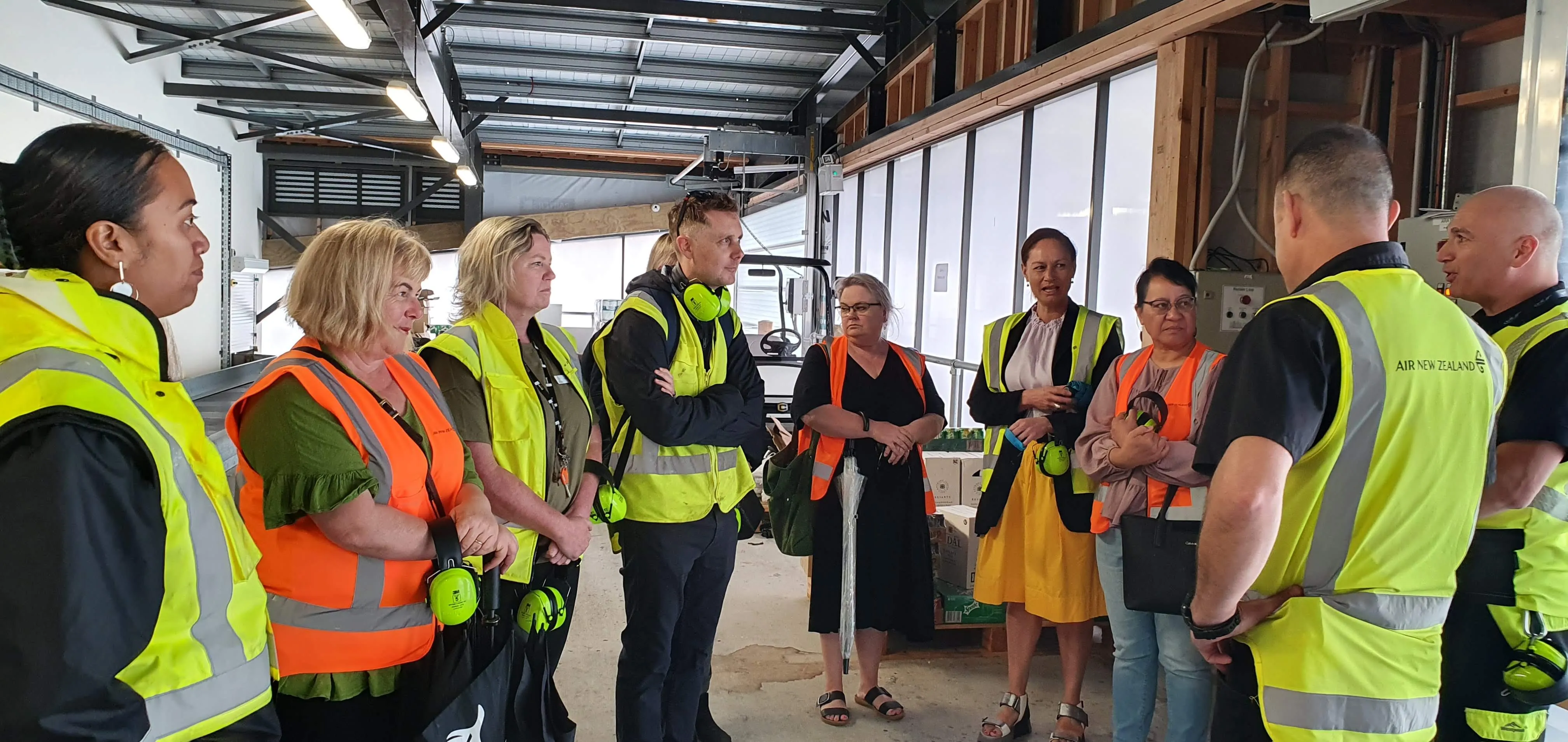 Group of people wearing high-visibility vests and holding safety earmuffs standing and talking inside a warehouse.