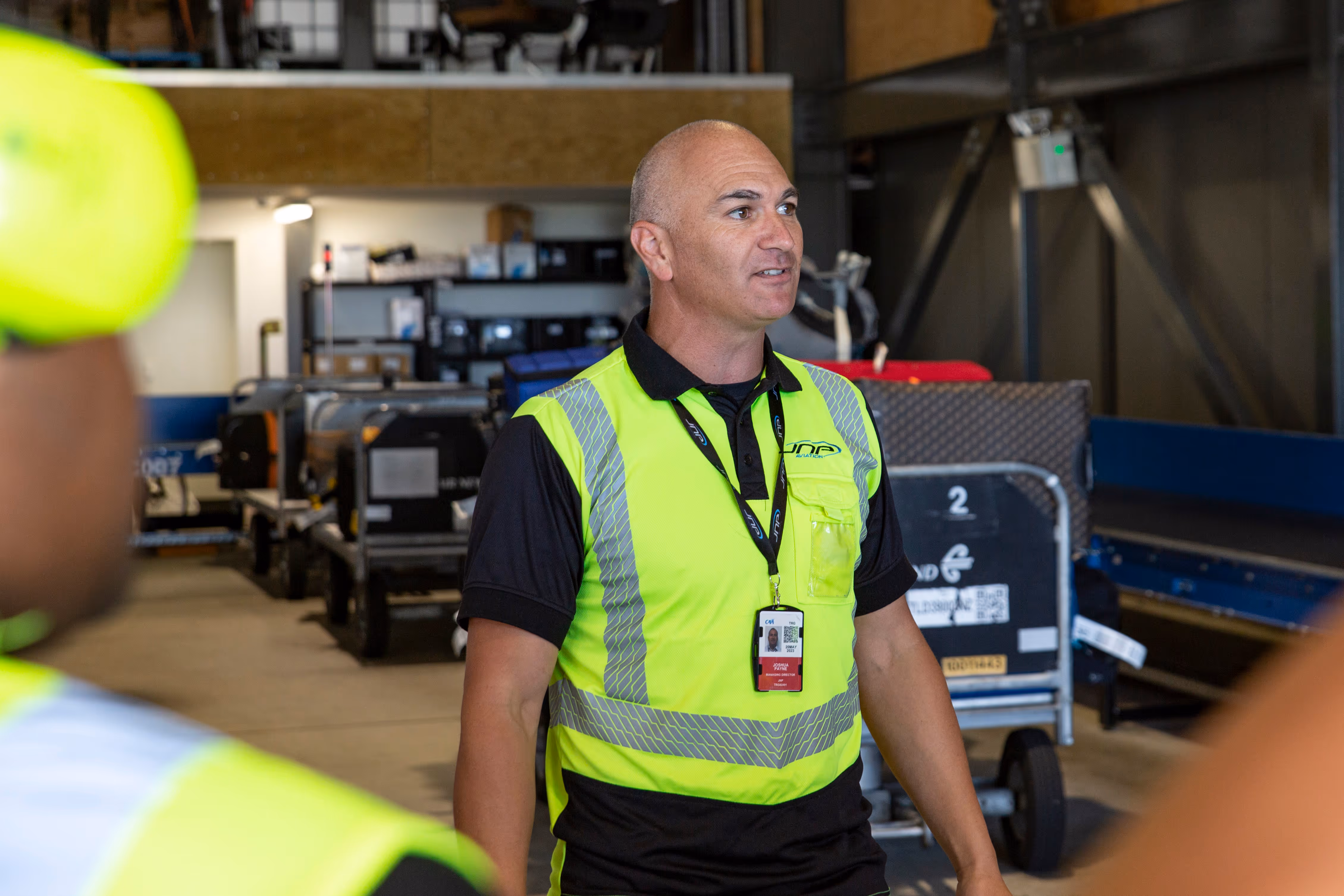 Man in a neon yellow safety vest speaking in an industrial setting with luggage carts in the background.