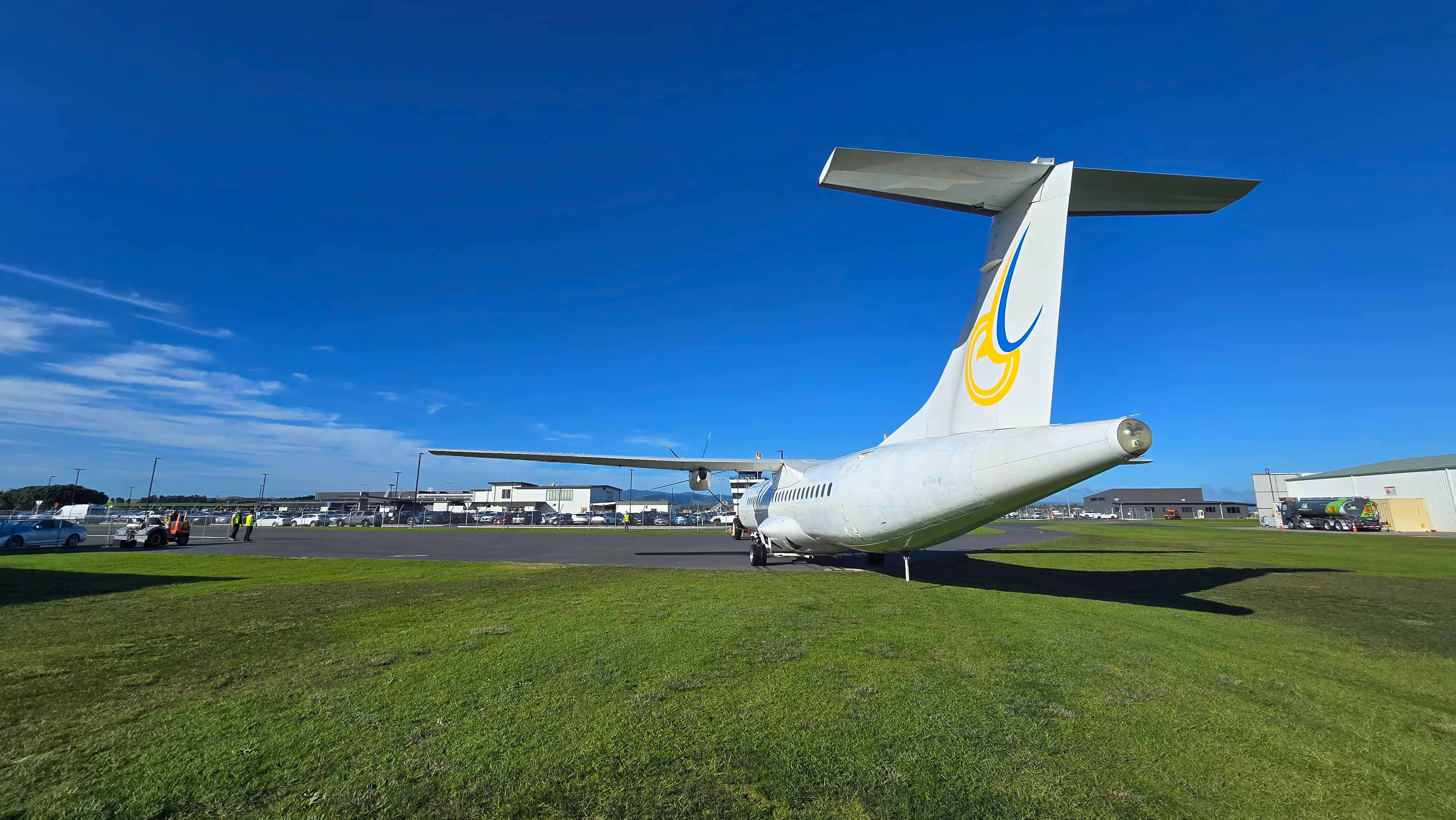 Small white airplane with yellow and blue logo parked on green grass near airport buildings under a clear blue sky.