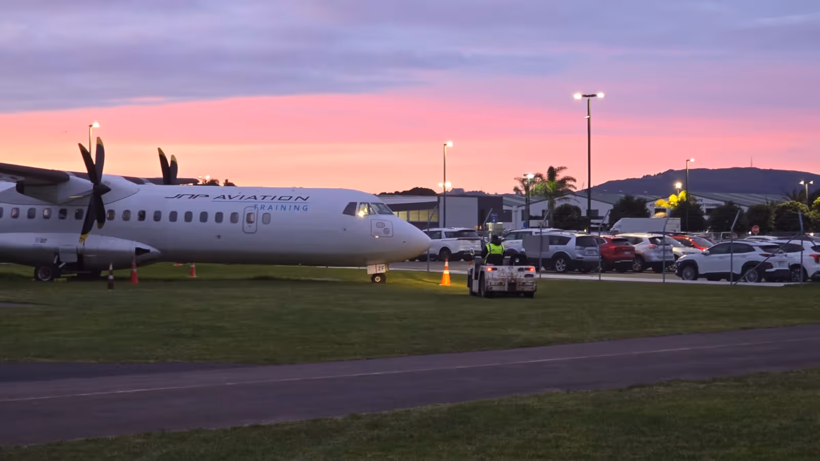 JMP Aviation training aircraft on grass near a parking lot at sunset with a ground vehicle nearby.