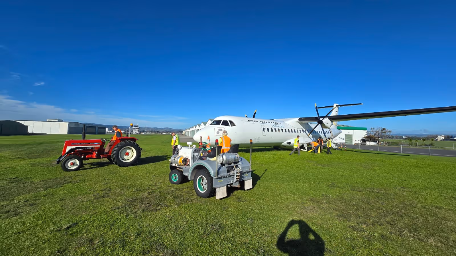 People in safety vests working around a white JNP Aviation Training propeller plane on grass near a red tractor and a small gray utility vehicle under a clear blue sky.