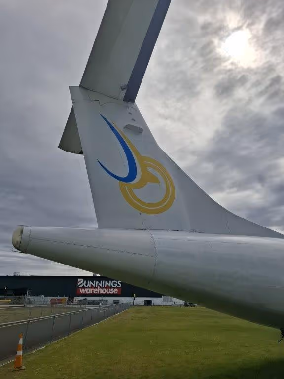 Close-up view of an airplane tail fin with a blue and yellow logo, under a cloudy sky with the sun faintly visible.