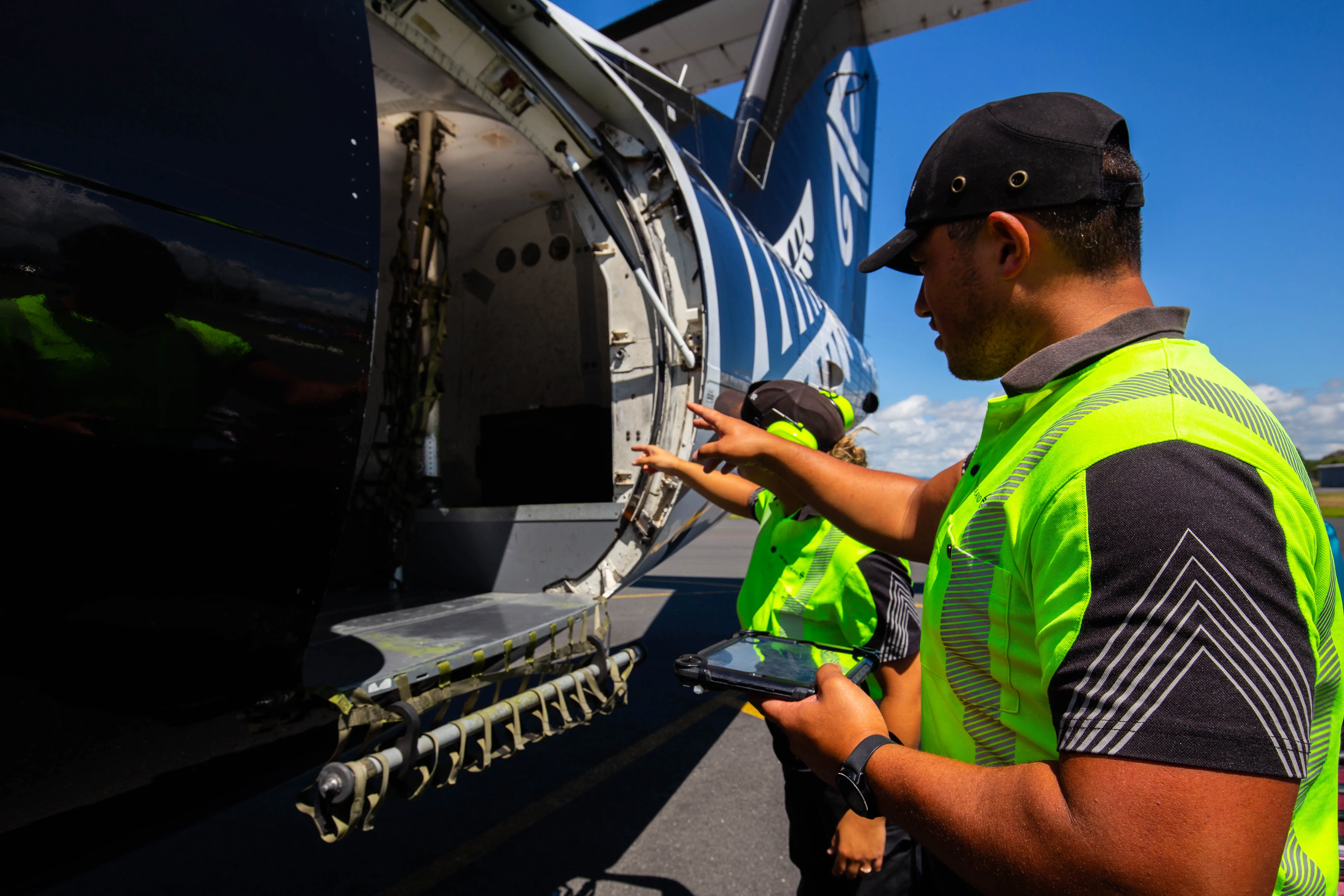 Two airport ground crew workers in neon yellow safety vests inspecting and pointing at the open cargo hold of an aircraft.