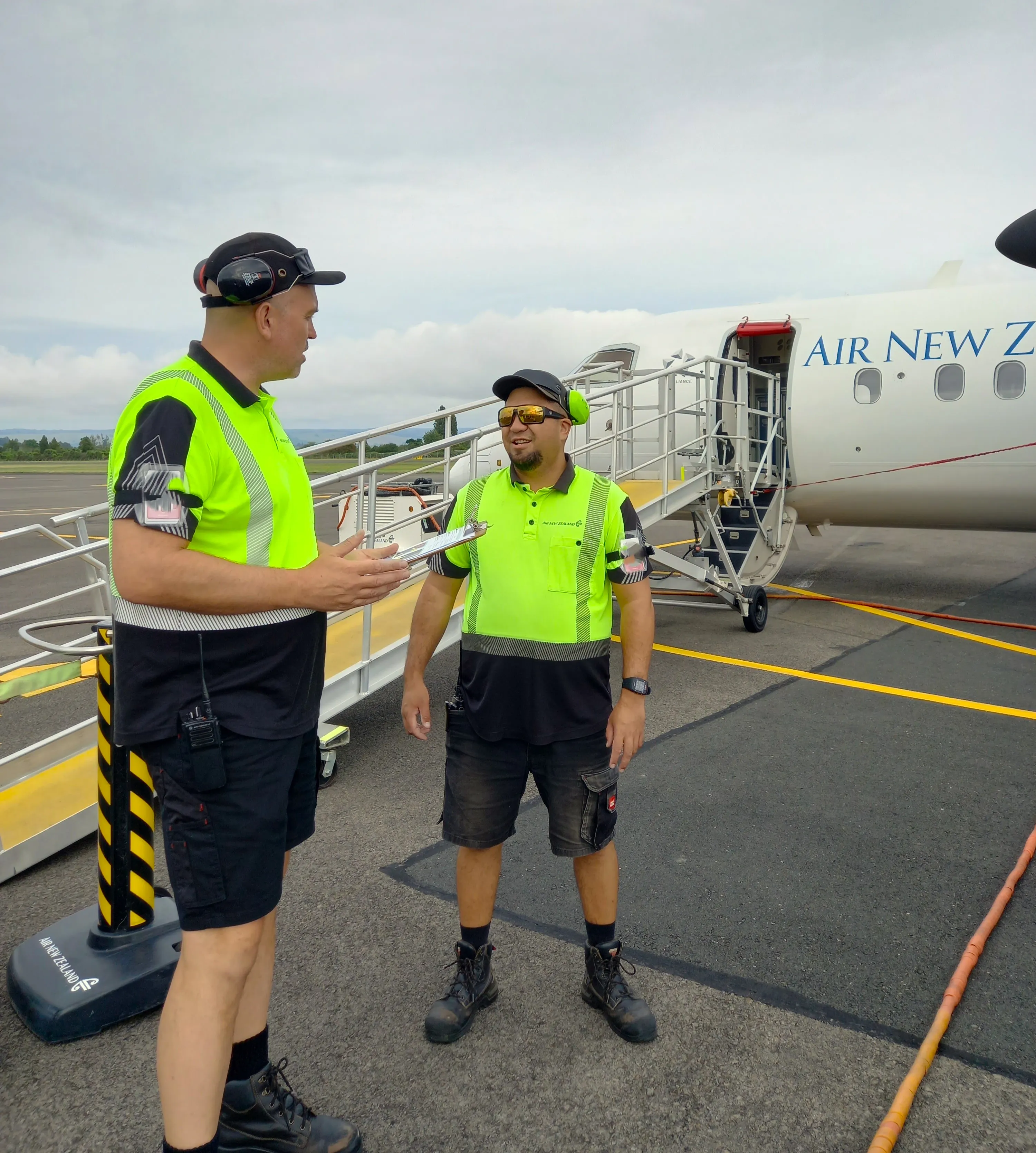 Two airport ground staff in high-visibility uniforms standing and talking near a stairway to an Air New Zealand plane.