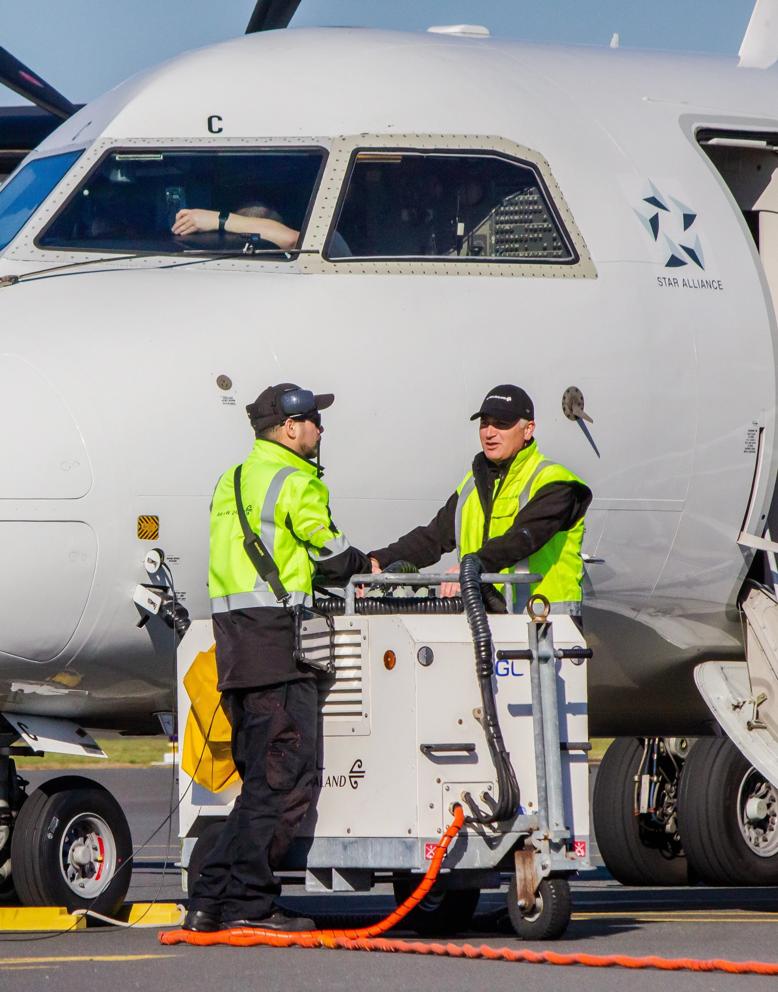 Two ground crew workers in yellow safety vests operating equipment near the nose of a commercial airplane with a pilot visible in the cockpit window.