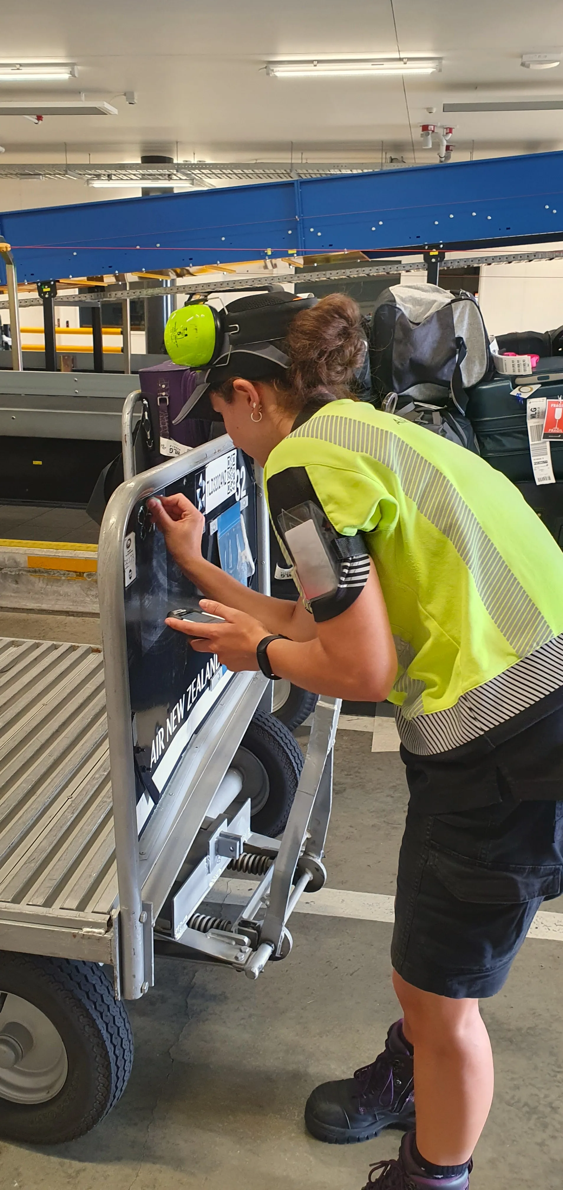 Female airport worker in a safety vest inspecting or scanning luggage on a baggage cart labeled Air New Zealand.