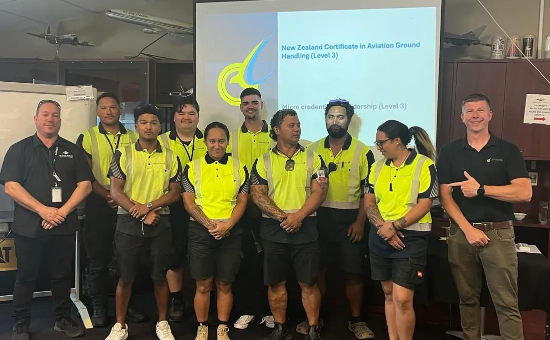 Group of nine people in high-visibility yellow shirts and two men in black shirts standing indoors in front of a screen displaying 'New Zealand Certificate in Aviation Ground Handling (Level 3)'.