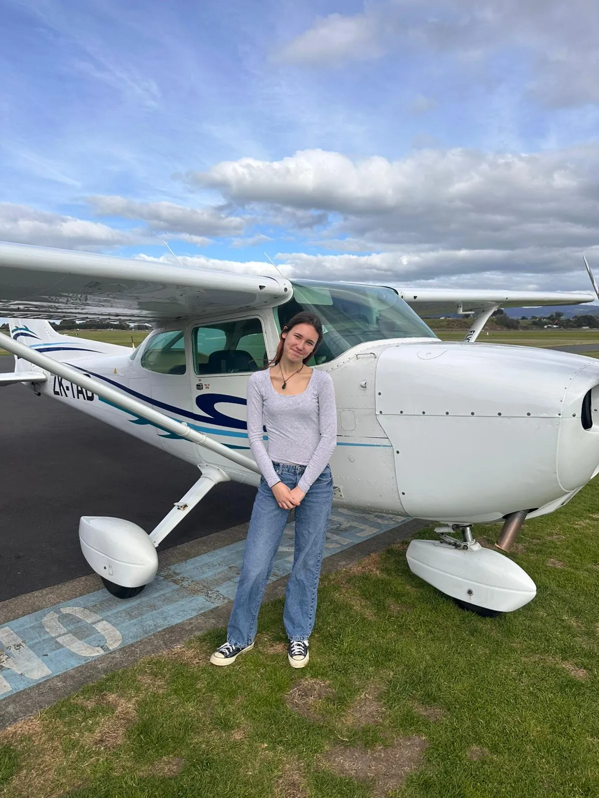 Young woman standing in front of a white small airplane on a grassy airfield under a cloudy sky.