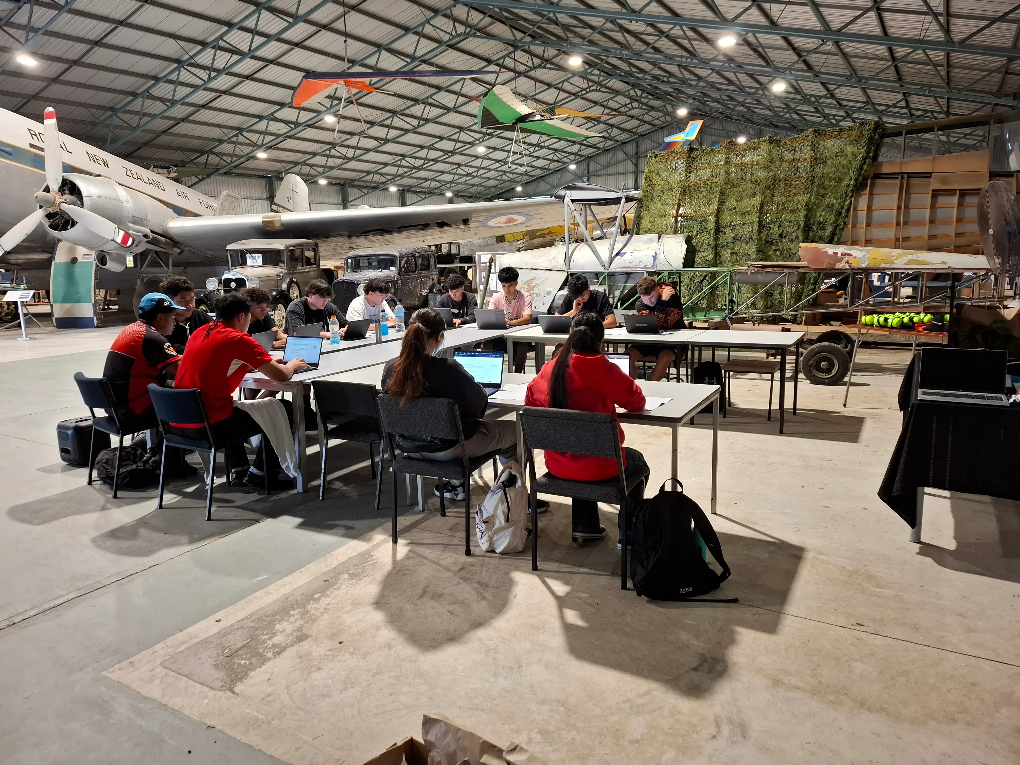 A group of people sitting around tables working on laptops inside an aircraft hangar with vintage planes and cars displayed in the background.