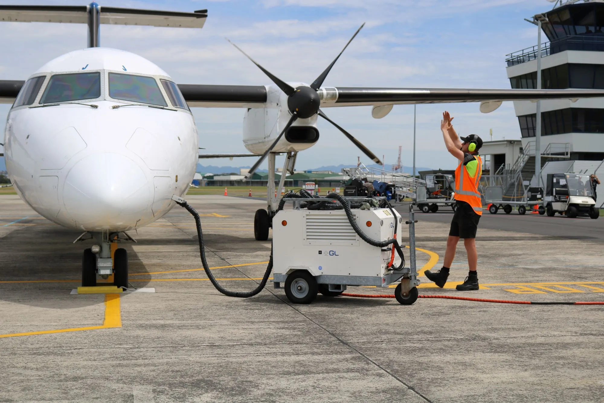 Marshalling worker in orange vest and ear protection guiding a white propeller airplane on an airport tarmac.
