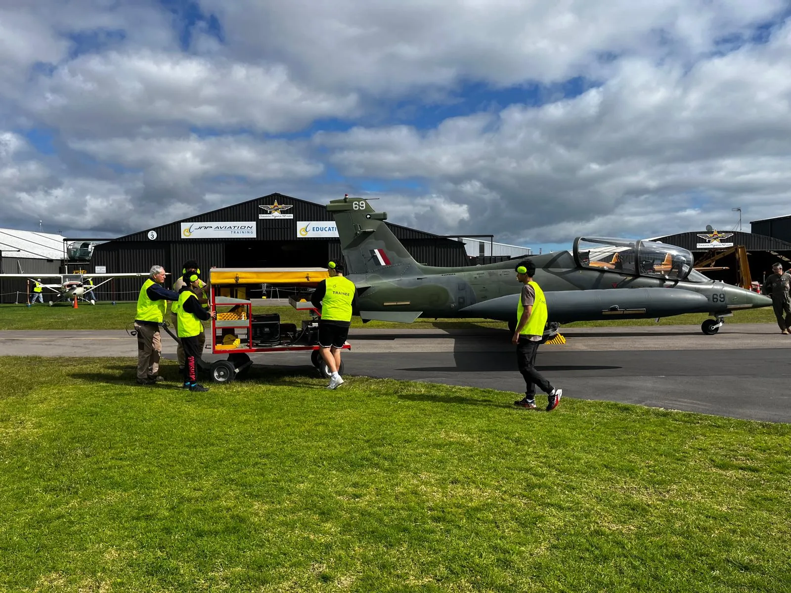 Four people in yellow safety vests and ear protection pushing equipment near a military jet aircraft parked on a runway with hangars in the background.