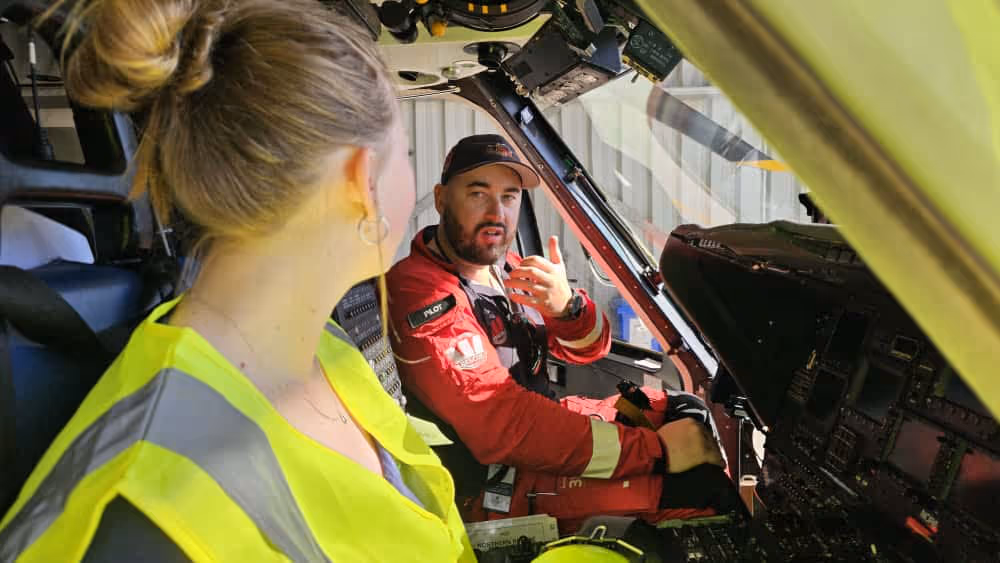 Pilot in red uniform and cap seated inside a cockpit speaking to a woman in a yellow safety vest.