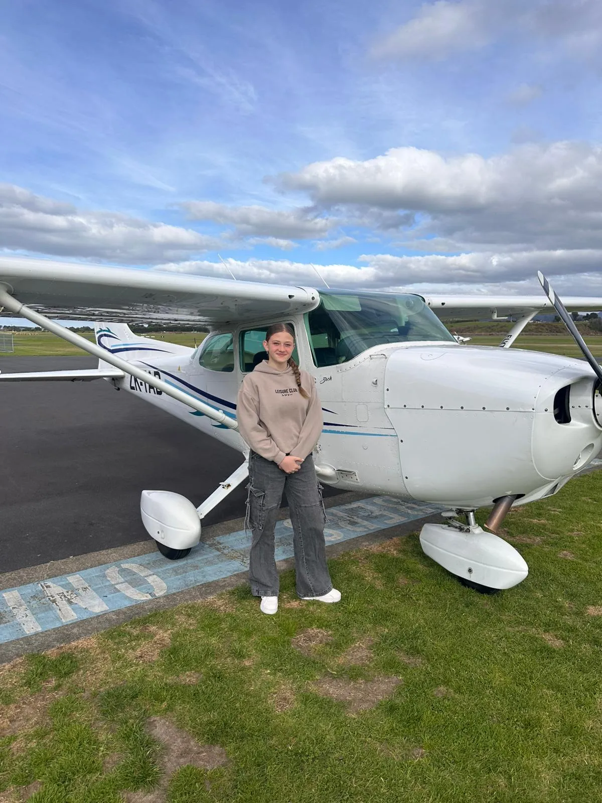 Young woman standing in front of a small white single-engine airplane on grass near a runway.
