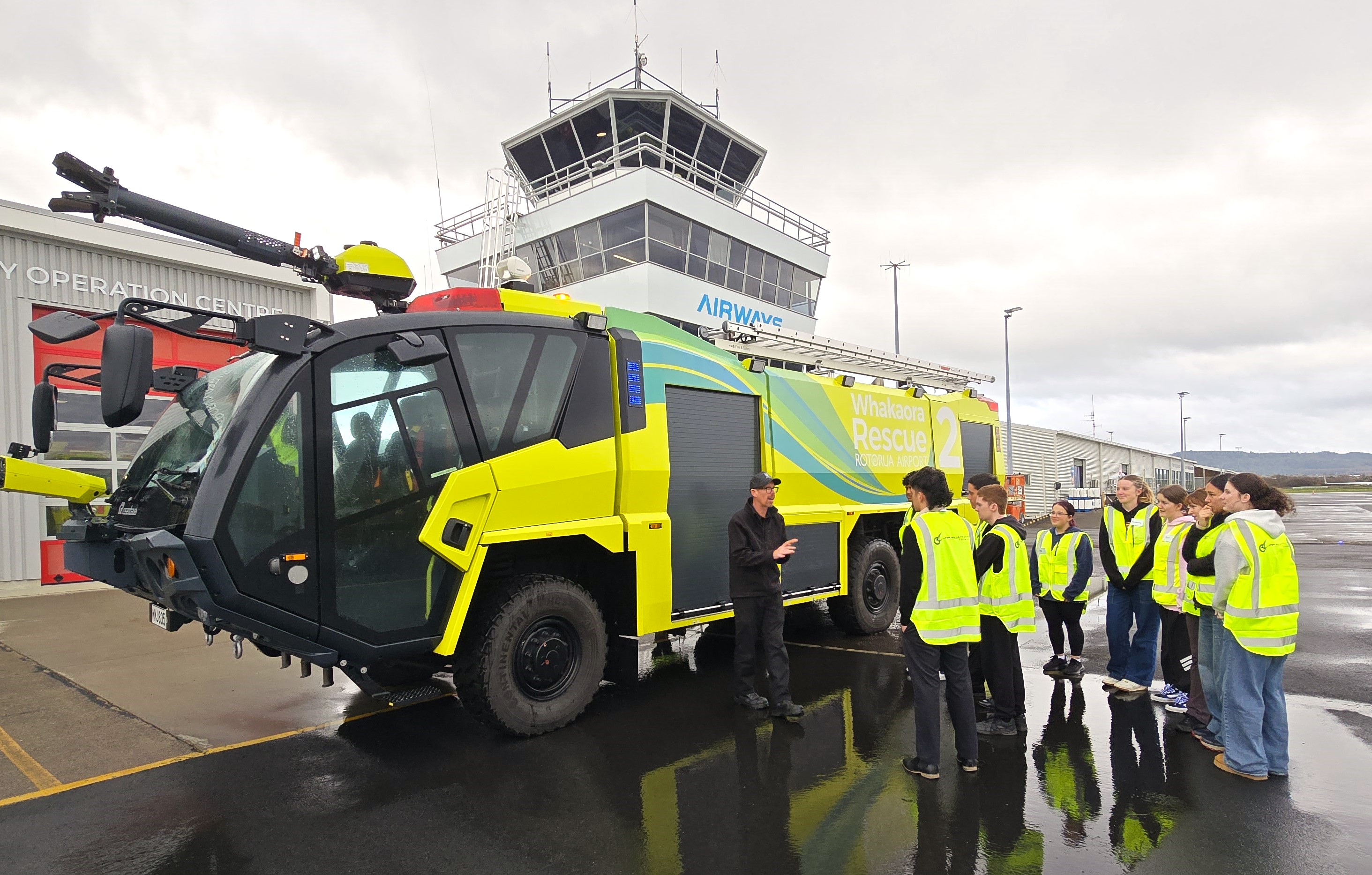 Group of people in yellow safety vests listening to a person explaining next to a large yellow airport rescue fire truck outside an airport control tower.