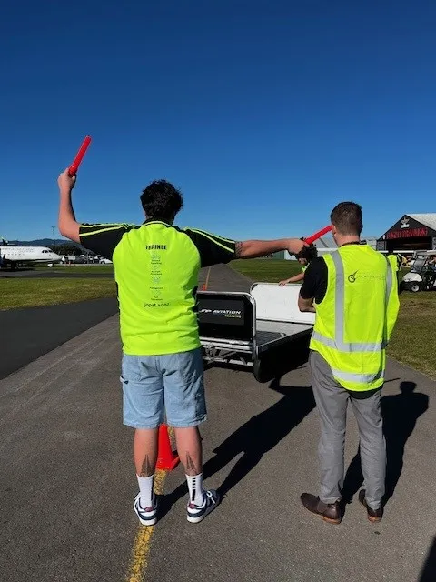 Two men wearing bright yellow reflective vests guide an aircraft on the tarmac using red signaling batons under a clear blue sky.