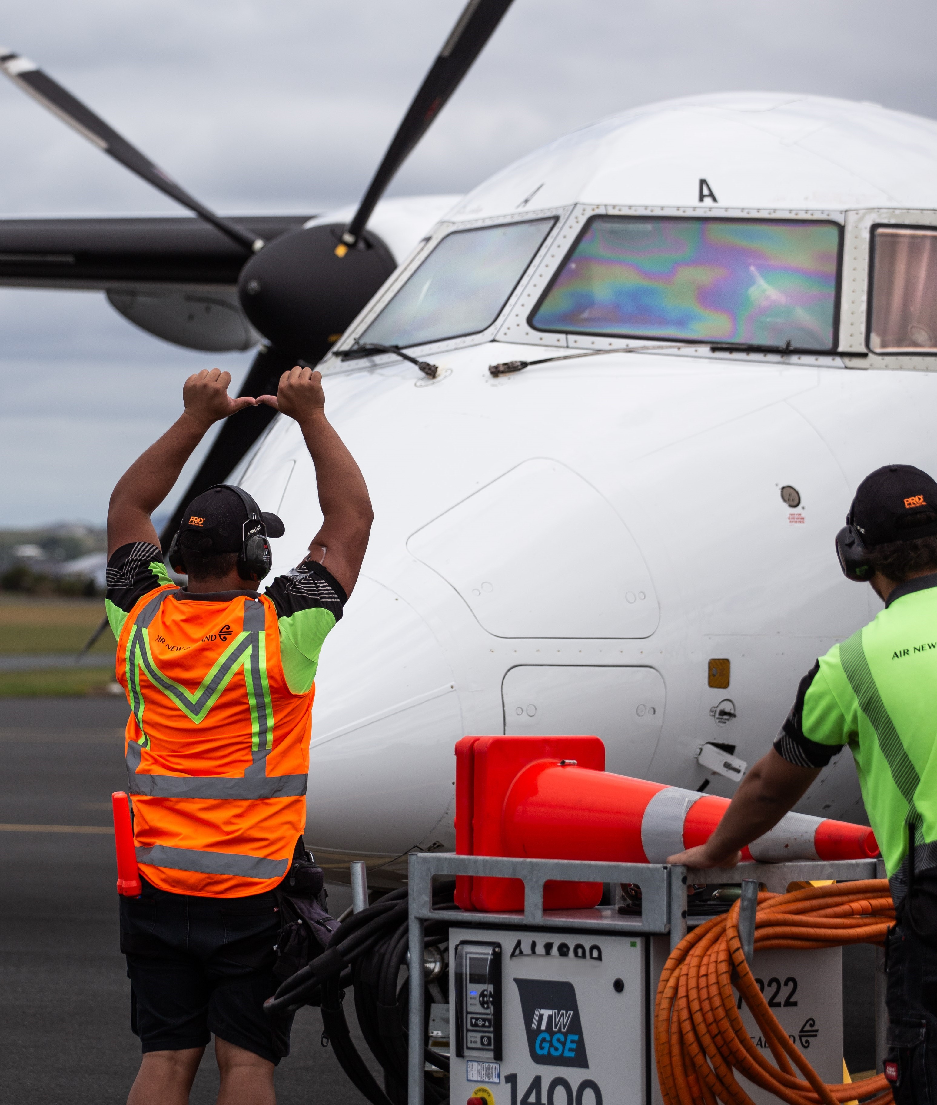 Ground crew in high-visibility vests guiding a white propeller airplane on the tarmac near equipment and orange traffic cones.