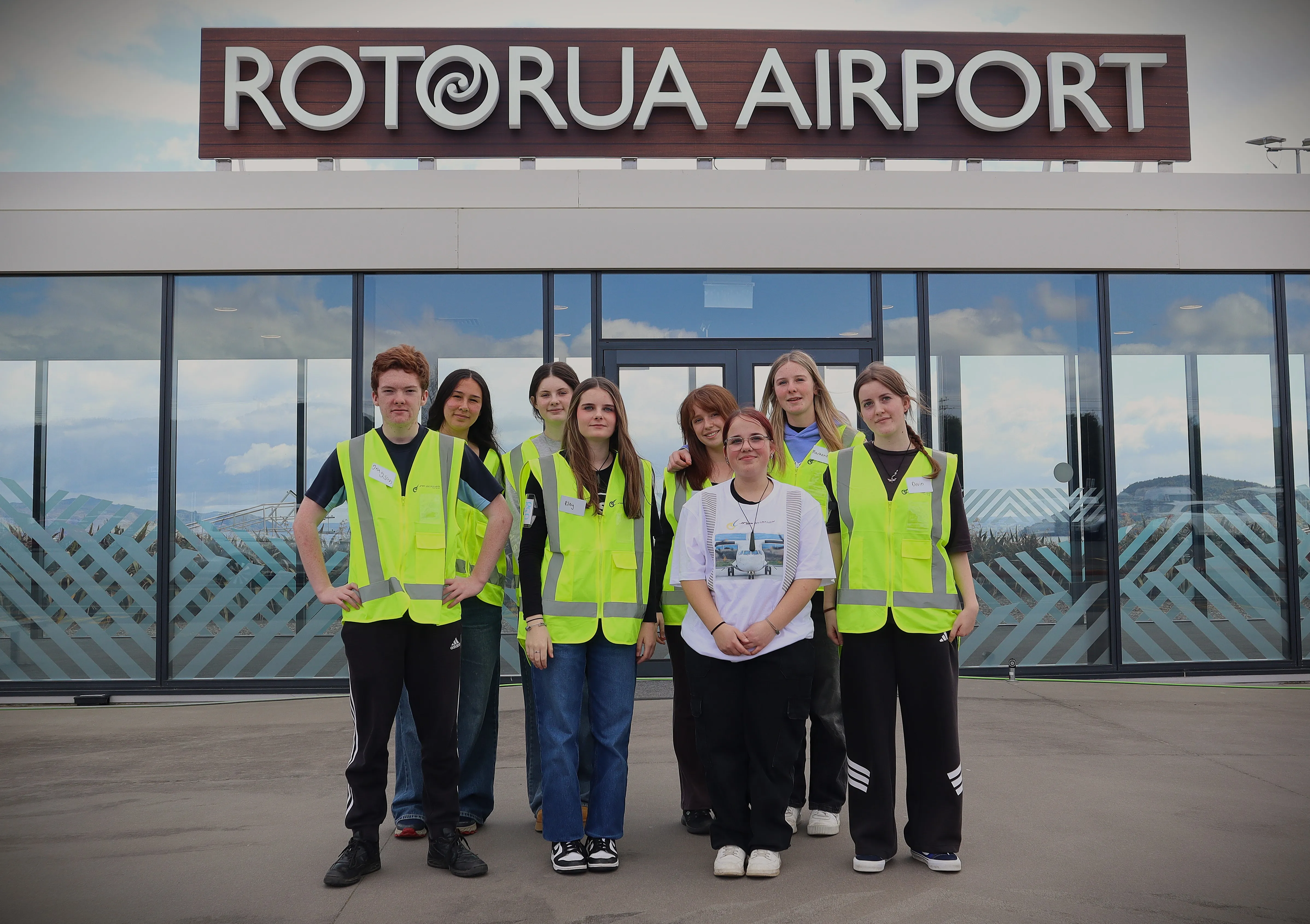 Group of seven young people wearing bright yellow safety vests posing outside Rotorua Airport in front of glass doors and large airport sign.