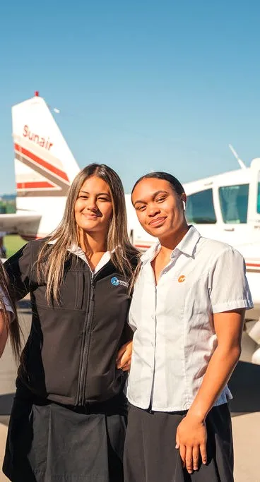 Two women smiling and posing in front of a small white airplane on a sunny day.