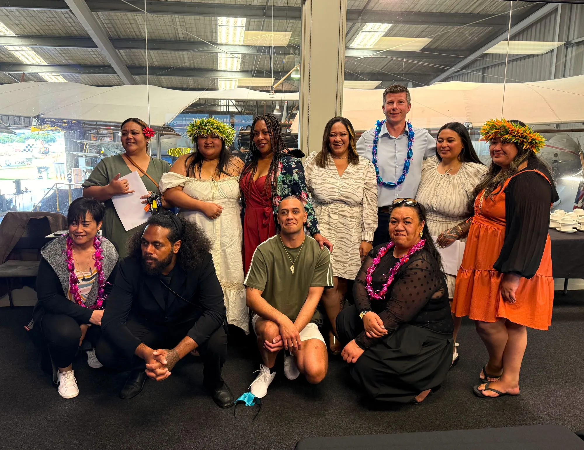 Group of thirteen people posing indoors, some wearing flower leis and traditional Polynesian attire, smiling at the camera.