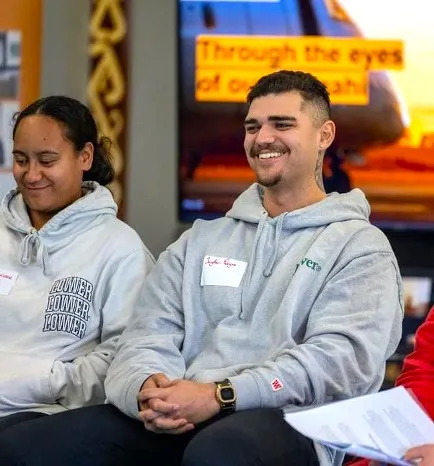 Two people sit side by side smiling, both wearing hoodies and name tags in an indoor setting.