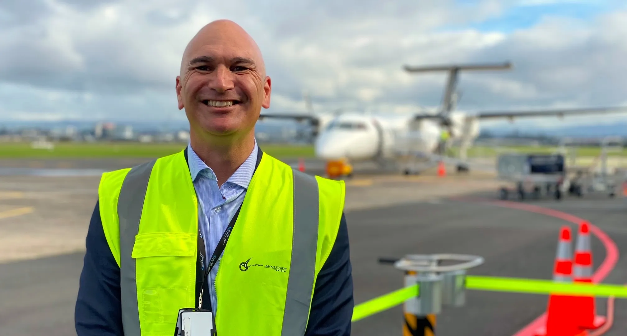 Smiling man wearing a high-visibility yellow vest standing on an airport tarmac with a small airplane and safety cones in the background.